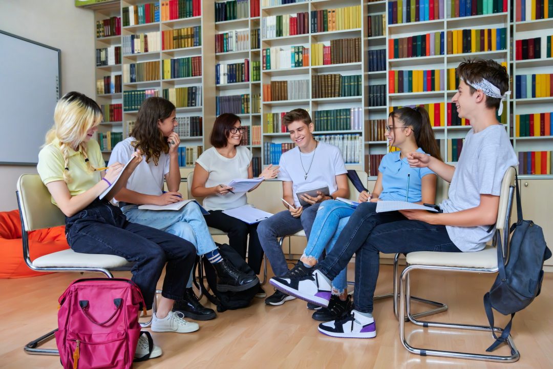 Obraz przedstawia grupę młodych osób siedzących na krzesłach w bibliotece, rozmawiających i notujących coś w zeszytach podczas spotkania.